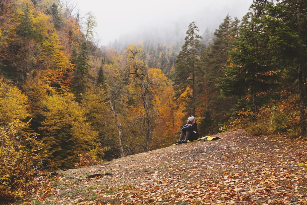 man in black jacket sitting on brown dirt road between green and brown trees during daytime