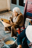 An elderly woman with white hair sits on a small red stool, wearing a brown outfit. She holds a woven basket that rests on her lap. Next to her, there is a stack of plastic chairs and a pot filled with food on the floor. The background shows decorative tiles and a wooden door.