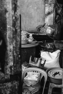 A cluttered indoor space with various objects including chairs, plastic bags, bowls, and a white sack. The wall features chalk-written numbers and words. The overall setting appears disorganized, with items stacked haphazardly.