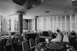 A black and white image of an empty banquet hall with several round tables arranged neatly. Each table is draped with a tablecloth and set with crockery. The room has a chandelier hanging from the ceiling, large windows with curtains, and panelled walls. A woman in a white dress is seated alone at one of the tables, occupied with a device.