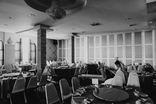 A black and white image of an empty banquet hall with several round tables arranged neatly. Each table is draped with a tablecloth and set with crockery. The room has a chandelier hanging from the ceiling, large windows with curtains, and panelled walls. A woman in a white dress is seated alone at one of the tables, occupied with a device.