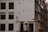 A street view of a corner building featuring a stark white facade with dark window frames and a number of windows. A classic street lamp is attached to the wall near the entrance, and a street sign with the text 'Binnenkant' is visible. Adjacent buildings are dark brown, and various architectural elements are clearly seen. The scene depicts a quiet, possibly residential area with a few bare trees visible.