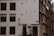 A street view of a corner building featuring a stark white facade with dark window frames and a number of windows. A classic street lamp is attached to the wall near the entrance, and a street sign with the text 'Binnenkant' is visible. Adjacent buildings are dark brown, and various architectural elements are clearly seen. The scene depicts a quiet, possibly residential area with a few bare trees visible.