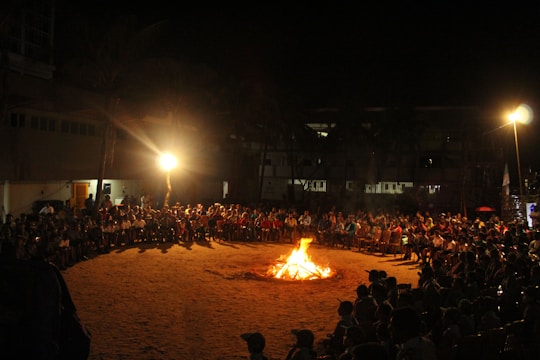 A large group of people sit in a circle around a bonfire on a sandy surface. The scene is illuminated by the fire and an overhead light, casting a warm glow on the gathering. The background consists of a building with dimly lit windows and palm trees, suggesting an outdoor community or festive event.