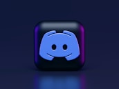 A 3D-style block with a blue logo in the center, set against a dark background. The logo resembles a game controller with two rounded edges and two circles on the front. The block has a subtle gradient, transitioning from dark to purple on its sides.