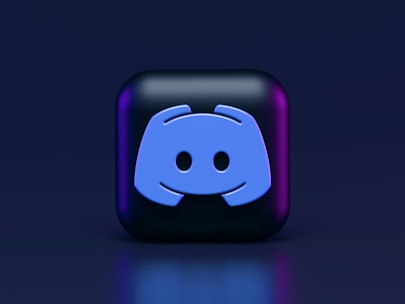 A 3D-style block with a blue logo in the center, set against a dark background. The logo resembles a game controller with two rounded edges and two circles on the front. The block has a subtle gradient, transitioning from dark to purple on its sides.
