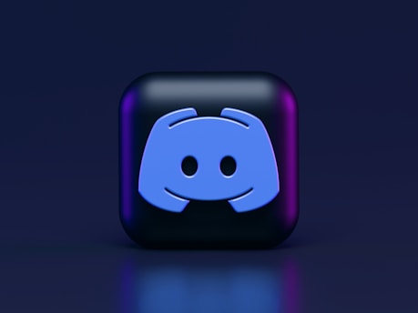 A 3D-style block with a blue logo in the center, set against a dark background. The logo resembles a game controller with two rounded edges and two circles on the front. The block has a subtle gradient, transitioning from dark to purple on its sides.