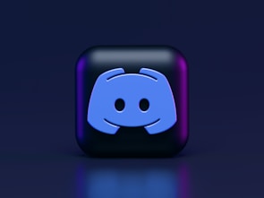 A 3D-style block with a blue logo in the center, set against a dark background. The logo resembles a game controller with two rounded edges and two circles on the front. The block has a subtle gradient, transitioning from dark to purple on its sides.
