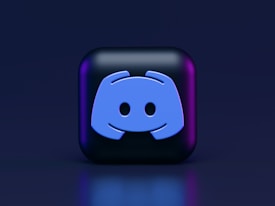 A 3D-style block with a blue logo in the center, set against a dark background. The logo resembles a game controller with two rounded edges and two circles on the front. The block has a subtle gradient, transitioning from dark to purple on its sides.