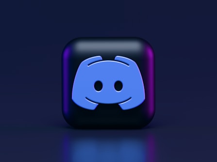 A 3D-style block with a blue logo in the center, set against a dark background. The logo resembles a game controller with two rounded edges and two circles on the front. The block has a subtle gradient, transitioning from dark to purple on its sides.