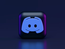 A 3D-style block with a blue logo in the center, set against a dark background. The logo resembles a game controller with two rounded edges and two circles on the front. The block has a subtle gradient, transitioning from dark to purple on its sides.