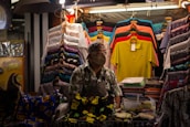 A person wearing a floral shirt and face mask stands behind a stall displaying a variety of colorful shirts and fabrics. The stall is well-lit with stacked and hanging shirts in different patterns and colors, including bright yellows, greens, reds, and blues.