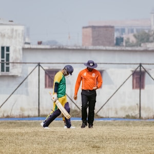 A cricketer in a green and yellow uniform walks alongside an umpire wearing an orange shirt and a navy blue hat. They are on a cricket field with a grassy surface. In the background, there is a wire fence and several buildings.