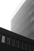 A monochrome photograph of modern architectural buildings showcasing a combination of geometric shapes and lines. The structure in the foreground features a series of vertical elements while the background building has a smooth facade with horizontal slats.