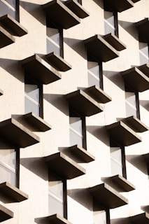 An architectural facade with a repetitive pattern of protruding rectangular balconies or window ledges casting shadows against a light-colored wall. The design features clean lines and uniform spacing, creating a geometric and modern aesthetic.