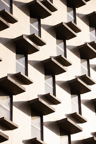 An architectural facade with a repetitive pattern of protruding rectangular balconies or window ledges casting shadows against a light-colored wall. The design features clean lines and uniform spacing, creating a geometric and modern aesthetic.