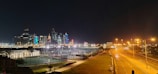 A nighttime cityscape with illuminated skyscrapers on the skyline. The foreground features a well-lit tennis court and a wide road with streetlights. The sky is dark, enhancing the brightness of the city lights.