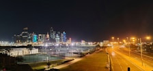 A nighttime cityscape with illuminated skyscrapers on the skyline. The foreground features a well-lit tennis court and a wide road with streetlights. The sky is dark, enhancing the brightness of the city lights.