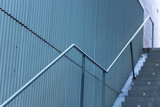 A corrugated metal wall runs alongside a set of concrete stairs that are ascending to the right. A metal handrail follows the incline of the stairs, contrasting against the uniform vertical lines of the wall.