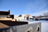 A partially blurred industrial scene featuring a long rusted metal beam in the foreground and several industrial buildings in the background under a clear blue sky. Snow is visible on the ground, and some structures are indistinct due to depth of field focus.