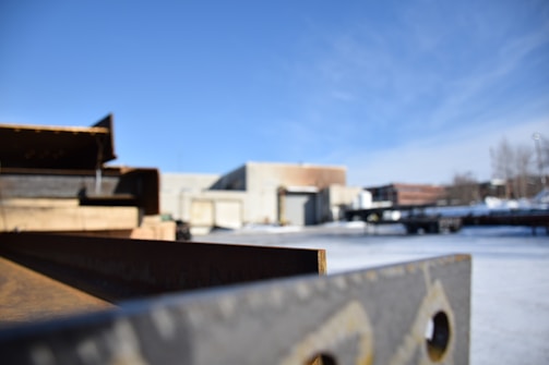 A partially blurred industrial scene featuring a long rusted metal beam in the foreground and several industrial buildings in the background under a clear blue sky. Snow is visible on the ground, and some structures are indistinct due to depth of field focus.