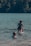 A group of people enjoys swimming and playing in a lake. One adult lifts a child in colorful swimwear above the water, while another child happily wades nearby. In the background, a dense forest borders the lake.