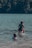 A group of people enjoys swimming and playing in a lake. One adult lifts a child in colorful swimwear above the water, while another child happily wades nearby. In the background, a dense forest borders the lake.