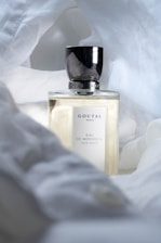 A glass bottle of Goutal Paris Eau de Monsieur eau de toilette is surrounded by soft white fabric. The bottle features a sleek, reflective silver cap and a minimalist label.