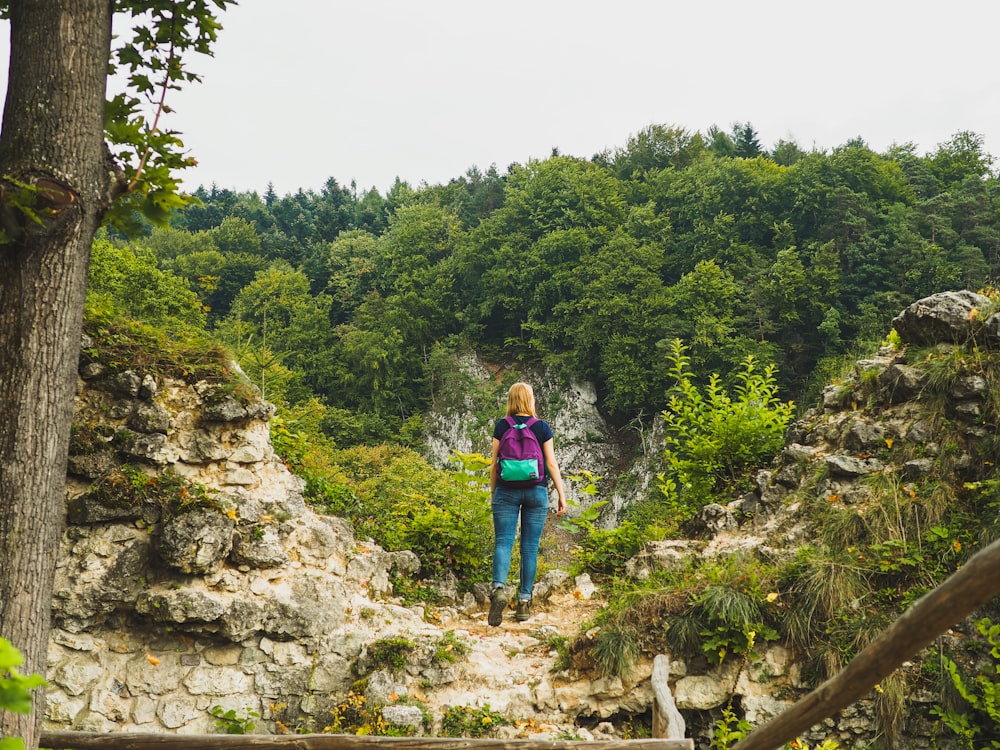 woman in blue jacket and blue backpack standing on rocky ground near green trees during daytime