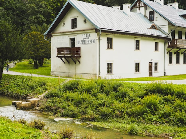 A rustic hotel building with a wooden balcony is set amidst lush greenery near a small stream. The structure has a pitched roof and multiple windows with a backdrop of dense trees. A path leads past the building, and a person is walking along it.