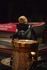 A close-up view of several objects on a dark surface. A stack of books, one titled 'Strategic Brand Management', is visible in the background. In the foreground, there's an ornate gold and patterned cup resting on a saucer. A small black sculpture, possibly depicting a mythical creature, is placed near the books, and on top of the sculpture is a light-colored snail shell.