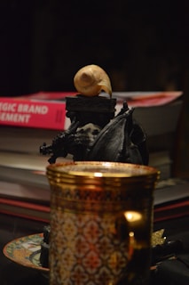 A close-up view of several objects on a dark surface. A stack of books, one titled 'Strategic Brand Management', is visible in the background. In the foreground, there's an ornate gold and patterned cup resting on a saucer. A small black sculpture, possibly depicting a mythical creature, is placed near the books, and on top of the sculpture is a light-colored snail shell.