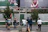 Several people are on a city street, with some performing juggling acts with colorful pins. Others stand around observing or walking past. In the background, large billboard advertisements are visible above a series of trees and urban structures.