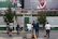 Several people are on a city street, with some performing juggling acts with colorful pins. Others stand around observing or walking past. In the background, large billboard advertisements are visible above a series of trees and urban structures.