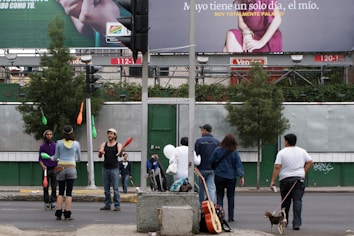 Several people are on a city street, with some performing juggling acts with colorful pins. Others stand around observing or walking past. In the background, large billboard advertisements are visible above a series of trees and urban structures.