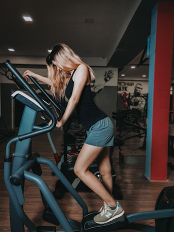 A woman with long hair is exercising on an elliptical machine in a gym environment. She is wearing a black tank top, grey shorts, and athletic shoes. The gym is dimly lit with visible workout equipment in the background, and there is a mural on the wall.