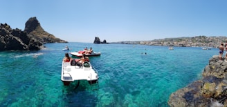 A scenic coastal view featuring clear blue water with several people enjoying activities. Some are on paddle boats, while others swim near rocky formations. The background showcases a distant town along the shoreline, with hills rising beyond. The sky is clear and bright.