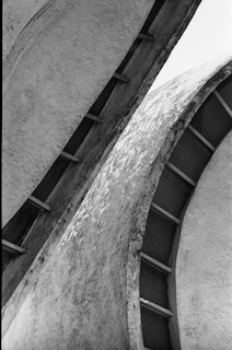 Curved, textured surfaces of a modern architectural structure create an abstract pattern with shadows and light contrasts. The image is composed of intersecting concrete forms with visible wear.