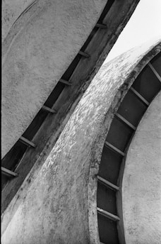 Curved, textured surfaces of a modern architectural structure create an abstract pattern with shadows and light contrasts. The image is composed of intersecting concrete forms with visible wear.