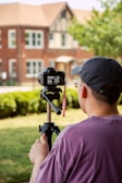 A person in a purple shirt and cap operates a camera mounted on a tripod. They are photographing or filming a brick house with architectural details in the background. The scene is outdoors, and there is a lawn with hedges and some trees.