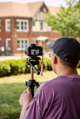 A person in a purple shirt and cap operates a camera mounted on a tripod. They are photographing or filming a brick house with architectural details in the background. The scene is outdoors, and there is a lawn with hedges and some trees.