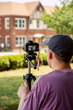 A person in a purple shirt and cap operates a camera mounted on a tripod. They are photographing or filming a brick house with architectural details in the background. The scene is outdoors, and there is a lawn with hedges and some trees.