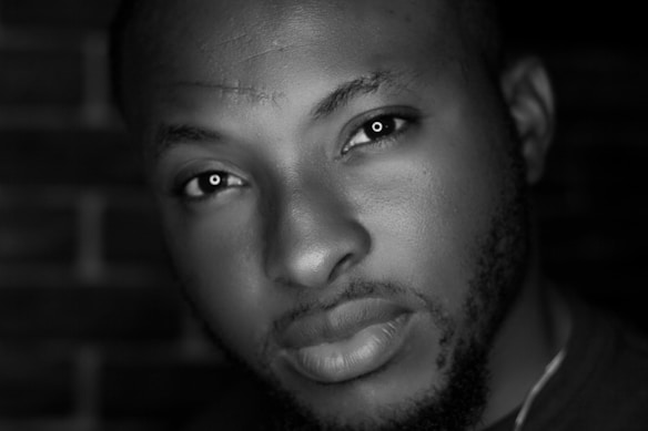 Close-up portrait of a person with a thoughtful expression in a black-and-white setting. The lighting highlights the facial features and texture of the skin against a blurred brick wall background.
