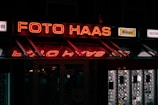 A neon sign reading 'Foto Haas' is prominently displayed, glowing in red against a dark background. Below the sign, there are reflections of the neon lights. On either side of the main sign, smaller advertisements for camera brands like Nikon and Sony are visible. The overall scene is dimly lit, suggesting it might be night, with additional signs and images visible inside a shop window.
