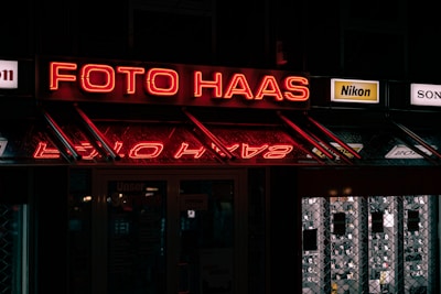 A neon sign reading 'Foto Haas' is prominently displayed, glowing in red against a dark background. Below the sign, there are reflections of the neon lights. On either side of the main sign, smaller advertisements for camera brands like Nikon and Sony are visible. The overall scene is dimly lit, suggesting it might be night, with additional signs and images visible inside a shop window.