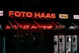 A neon sign reading 'Foto Haas' is prominently displayed, glowing in red against a dark background. Below the sign, there are reflections of the neon lights. On either side of the main sign, smaller advertisements for camera brands like Nikon and Sony are visible. The overall scene is dimly lit, suggesting it might be night, with additional signs and images visible inside a shop window.