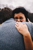 A close-up view of two people embracing, with a focus on their upper bodies. One person is wearing a textured blue sweater, and the other person, with short dark hair, has their eyes closed and is smiling contentedly. A hand with a ring rests gently on the back of the person in the sweater.