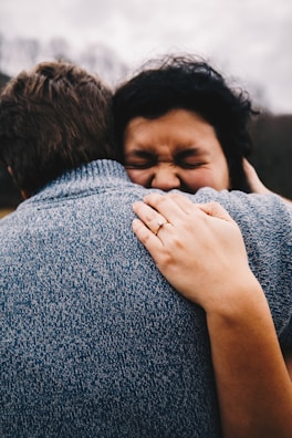 A close-up view of two people embracing, with a focus on their upper bodies. One person is wearing a textured blue sweater, and the other person, with short dark hair, has their eyes closed and is smiling contentedly. A hand with a ring rests gently on the back of the person in the sweater.