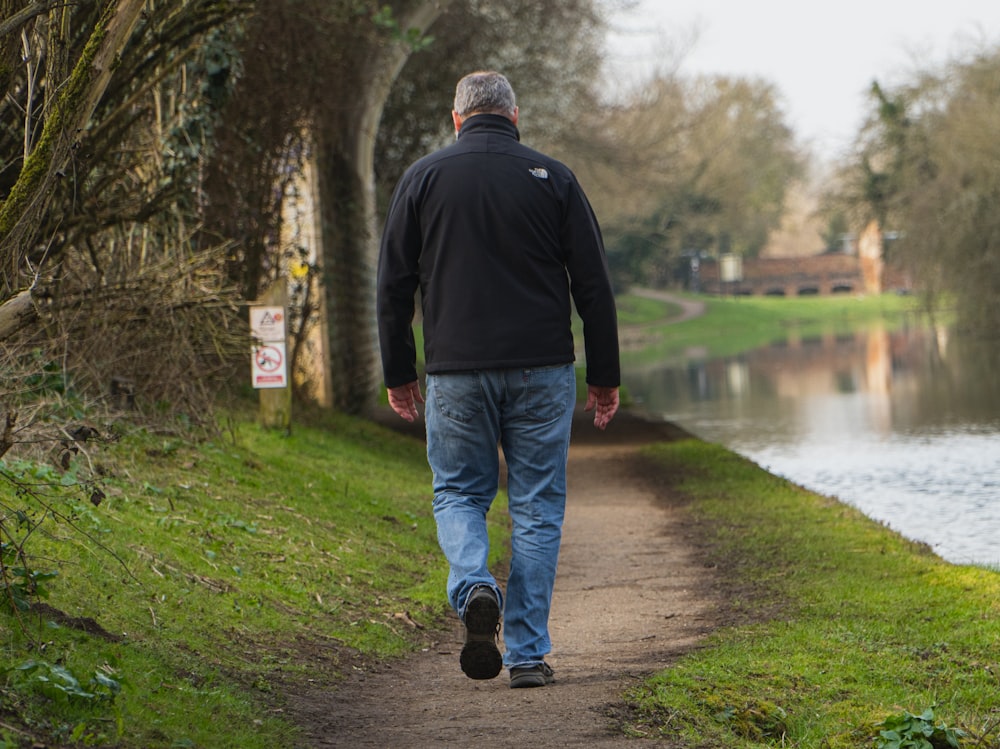 man in black jacket and blue denim jeans walking on pathway near river during daytime