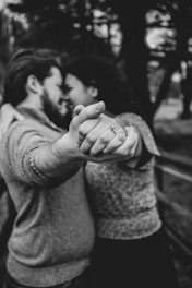 A couple is embracing closely with their faces nearly touching while the focus is on their intertwined hands in the foreground. One of them is wearing a ring, suggesting engagement or marriage. The scene is set outdoors with blurred trees in the background, captured in black and white.